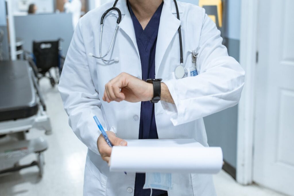 Doctor in lab coat checking wristwatch while holding medical chart in hospital corridor.